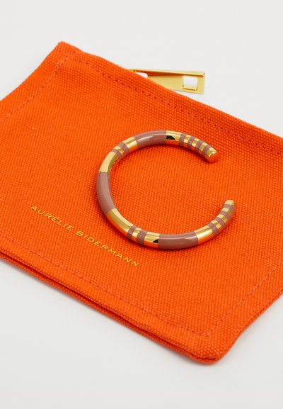 Bracciale marrone con strisce dorate su un sacchetto di tessuto arancione con testo in rilievo, caratterizzato da una finitura liscia e un design curvato.