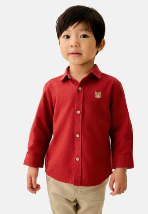 Junge mit kurzem dunklem Haar, der ein rotes Hemd mit Knöpfen und einem kleinen Bären-Patch sowie beige Hosen vor einfachem Hintergrund trägt.