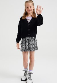 Cardigan nero a coste con tre bottoni, indossato sopra una maglietta viola e una gonna plissettata a zig-zag. Stivali bianchi con lacci a righe.