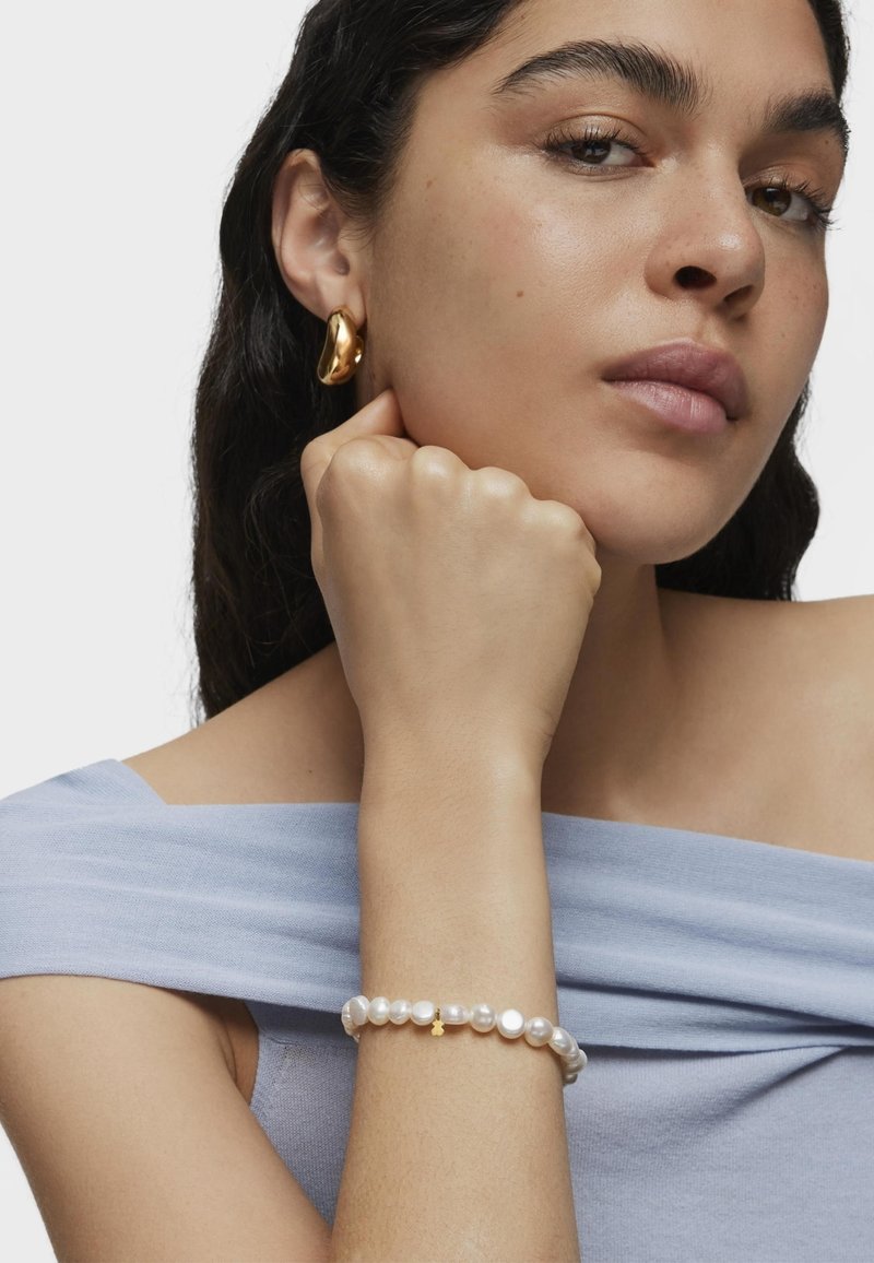 Pulsera de perlas con un acento dorado, que presenta una textura suave y perlas redondeadas y brillantes. El modelo lleva una blusa azul clara sin mangas.