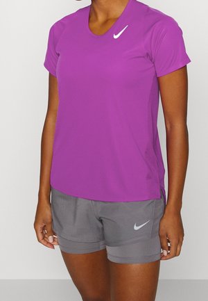 Kvinde iført en lilla Nike kortærmet skjorte og grå Nike sportsshorts, der står mod en ensfarvet baggrund.