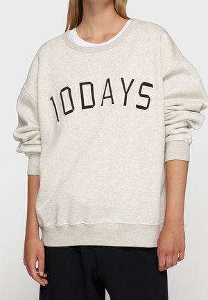 Personne portant un sweat-shirt beige clair avec le texte "10DAYS" en grandes lettres noires sur la poitrine et un pantalon noir.