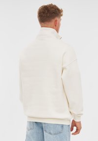 White high-collar sweatshirt with a relaxed fit and ribbed cuffs, paired with light blue denim jeans. Minimalistic design, soft texture.