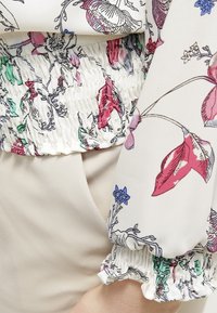 Blus med blommönster, smockad midja och elastiska ärmar. Har en krämfärgad botten med rosa, gröna och blå accenter; mjuk textur.