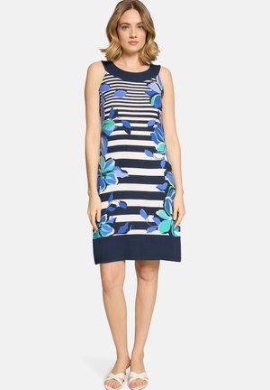 Frau trägt ärmelloses marineblau-weiß gestreiftes Kleid mit großem blauem Blumenmuster und weißen Sandalen, steht vor einfarbigem Hintergrund.