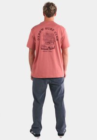 Camiseta de algodón rosa con un gráfico de "Oxbow Surf Club" y diseño de camión en la parte posterior. Combinada con pantalones azul oscuro y zapatillas negras.