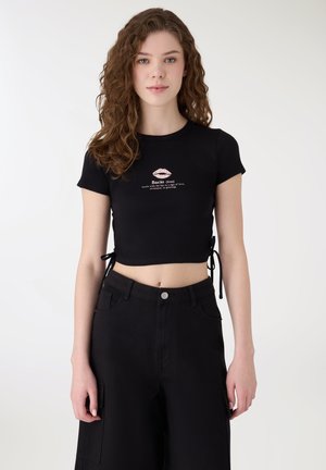 Giovane donna con capelli ricci che indossa un top corto nero con grafica di labbra e testo, abbinato a pantaloni neri a vita alta su sfondo semplice.