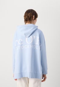 Hellblauer übergroßer Hoodie mit weicher Textur, der auf der Rückseite weißen Aufdruck mit dem Text "rc O' STOCKHOLM" und eine Kapuze mit Kordelzug aufweist.