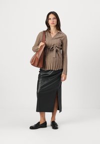 Brauner gerippter Strickpullover mit gebundenem Bund, kombiniert mit einem schwarzen Lederrock mit hohem Schlitz und schwarzen Loafers, während eine braune Schultertasche gehalten wird.