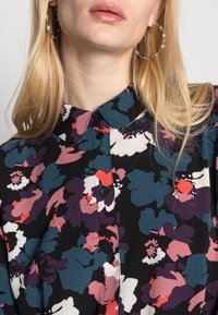 Blouse à fleurs à col en noir avec un mélange vibrant de violet, rouge et bleu. Comprend des boutons et un tissu texturé avec une coupe ample.
