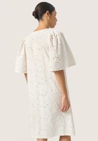Valkoinen mekko, jossa on lyhyet puff-hihat, ja se sisältää teksturoitua kangasta ja rei'itettyjä kuvioita. Yli polven pituus ja rento istuvuus.