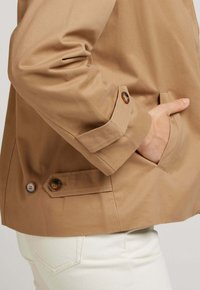 Veste en coton beige à la coupe décontractée, avec une poche et des accents de boutons sur les poignets des manches. Portée par-dessus un pantalon clair.