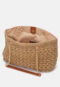 Beige pletená taška typu tote s texturovaným vzorem, zlatým řetězem a koženými akcenty, a měkkým béžovým podšívkou. Otevřený vrch se zapínáním na patentku.