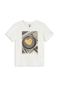 White cotton t-shirt featuring a printed graphic of a heart-shaped pancake in a frying pan, with a gray stove background.