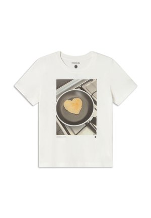 Weißes Baumwoll-T-Shirt mit einer bedruckten Grafik eines herzförmigen Pfannkuchens in einer Bratpfanne, vor einem grauen Herd-Hintergrund.