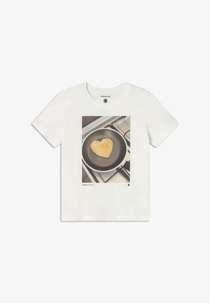 Biała bawełniana koszulka z nadrukiem graficznym przedstawiającym naleśnika w kształcie serca na patelni, z szarym tłem kuchennej kuchenki.