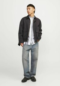 Giubbotto bomber nero con tasche frontali, indossato su una camicia a righe leggera con bottoni. I jeans blu larghi e le scarpe nere completano l'outfit.