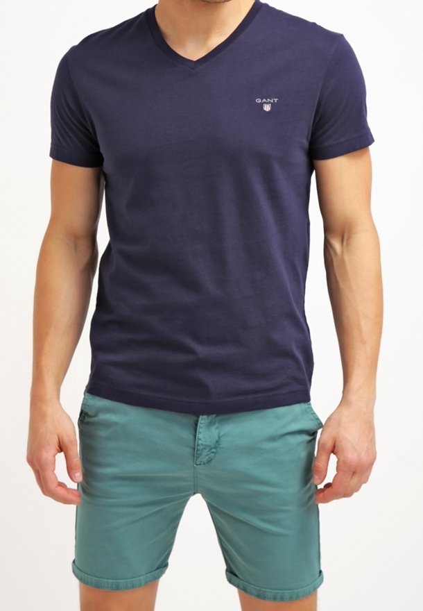 Homme portant un t-shirt bleu marine à col en V et un short turquoise retroussé, debout avec les mains détendues le long du corps.