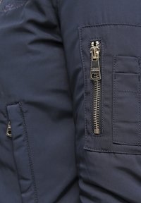 Donkerblauwe pufferjack met een gestructureerde stof, voorzien van een opvallende gouden rits op de linkermouw en een patchzak erboven.