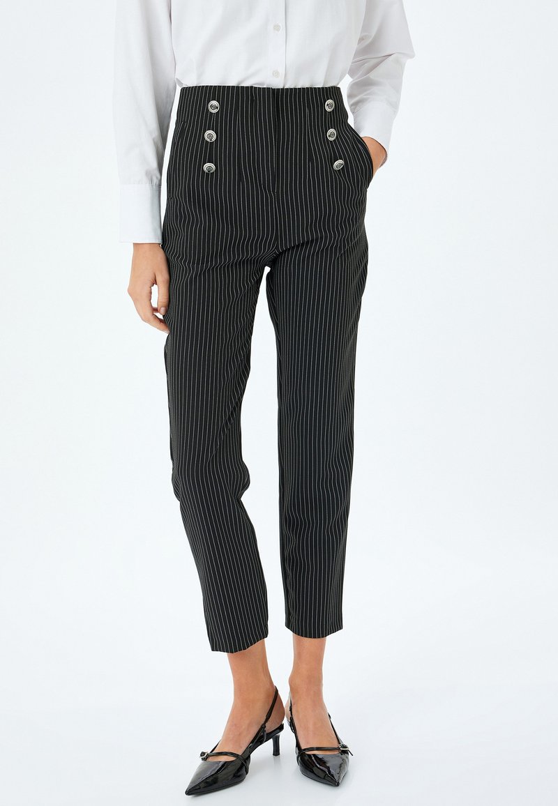Schwarze Nadelstreifenhose mit hoher Taille, versehen mit silbernen Knopfakzenten, vorderen Taschen und einem schmal zulaufenden Beindesign. Kombiniert mit schwarzen Absätzen.