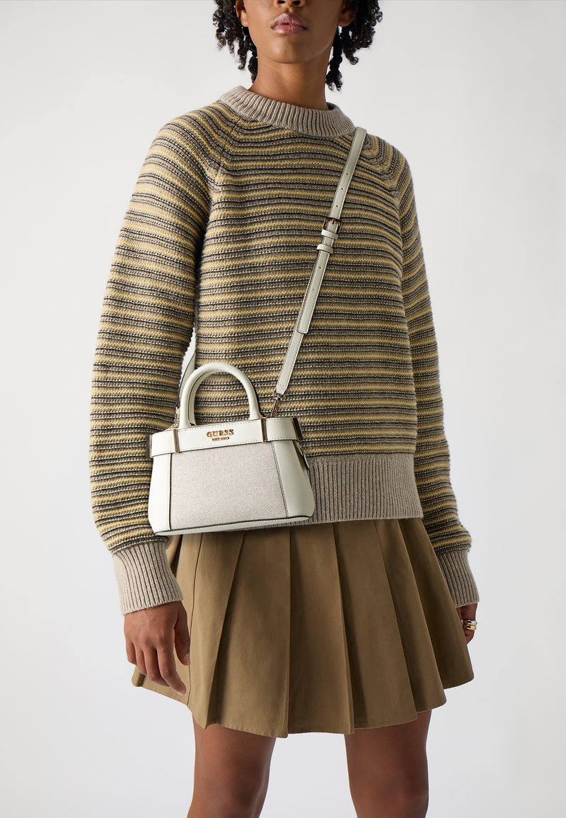 Kleine witte handtas met gouden logo, gestructureerde vorm, korte handvatten en een afneembare strap. Draagt bij een gestreepte trui en een bruine geplooide rok.