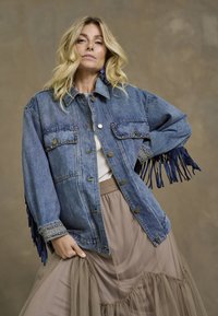 Jasnoniebieska kurtka dżinsowa z frędzlami przy mankietach, z ozdobnymi złotymi ćwiekami, zapinana na guziki, z dwiema kieszeniami na piersi, noszona na jasnej bluzce i beżowej spódnicy.