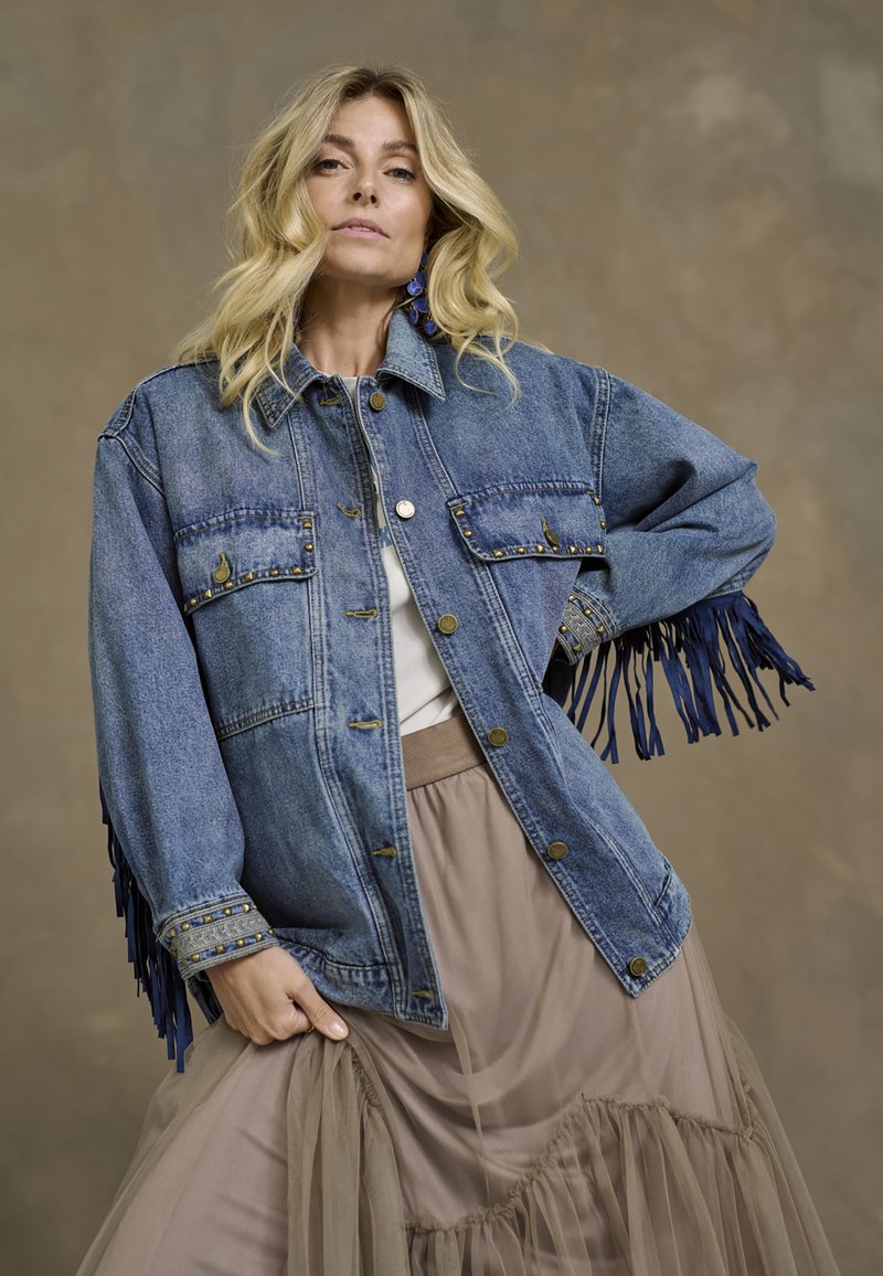 Jasnoniebieska kurtka dżinsowa z frędzlami przy mankietach, z ozdobnymi złotymi ćwiekami, zapinana na guziki, z dwiema kieszeniami na piersi, noszona na jasnej bluzce i beżowej spódnicy.