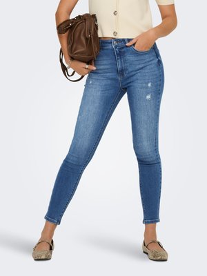 Femeie purtând blugi skinny albaștri cu talie înaltă și uzură minoră, top bej cu mâneci scurte, balerini din piele de șarpe și ținând o geantă maro din piele.