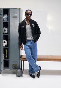 Man in sunglasses leaning against a bench beside an open locker with soccer balls, trophies, and a gray basketball on the floor.