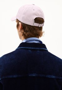 Helle rosa Baseballmütze mit verstellbarem Riemen, umgedreht getragen; blau gestreiftes Hemd sichtbar unter einer strukturierten dunkelblauen Cordjacke.