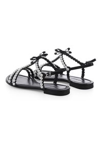 Schwarze flache Sandalen mit geflochtenem Design, verziert mit weißen Kunstperlen und schwarzen Schleifen. Mit einem Knöchelriemen und glatter Sohle.