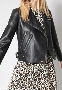 Mustasta nahasta valmistettu moottoripyörätakki, jossa on epäsymmetrinen vetoketju, useita vetoketjullisia taskuja ja vyö vyötäröllä. Kuvioitu mekko näkyy takin alta.