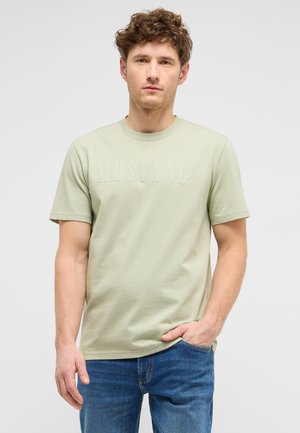 Junger Mann mit lockigem Haar, der ein hellgrünes "MUSTANG" T-Shirt und blaue Jeans trägt, steht mit einer Hand in der Tasche vor einem einfarbigen Hintergrund.