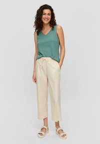 Haut sans manches en teal avec un col en V, associé à un pantalon crème à taille élastique et des sandales à lanières de couleur claire sur un mannequin.