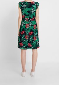 Nainen seisoo selkä kameraan, päällään vihreä trooppinen kukkakuosiset mekko ja valkoiset tennarit, taustanaan yksinkertainen tausta.