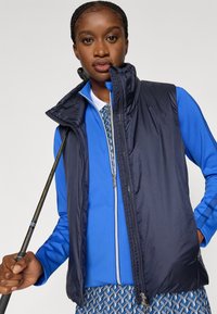 Colete azul-marinho sem mangas com gola alta, fecho frontal com zíper, combinado com uma camisa azul de manga comprida com padrão geométrico. Segura um taco de golfe na mão.