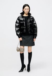 Μαύρο γυαλιστερό μπουφάν με καπιτονέ σχέδιο και κοντή γραμμή, μαύρο πλεκτό πουλόβερ, γκρίζα φούστα, μίνι μαύρες κάλτσες μέχρι το γόνατο και μια μικρή τσάντα με σχέδιο.