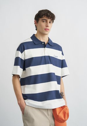 Noor mees seljas sinise ja valge triibulise polo-särgi ning beežide pükstega, vasakus käes oranži nokamütsu hoides, taust on ühevärviline.