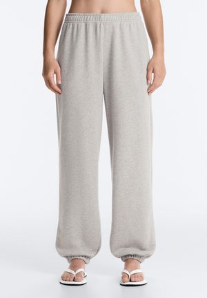 Personne portant un pantalon de survêtement gris clair à taille élastique avec poignets élastiques et des tongs blanches, debout devant un fond blanc uni.