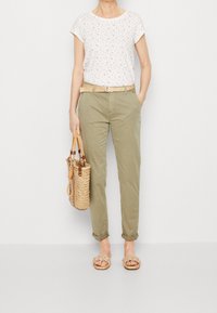 Bílý tričko s květinovým vzorem, olivově zelené kalhoty, tkaný opasek, slámová kabelka a hnědé pletené sandály. Neformální a pohodlný outfit.