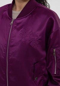 Paarse satijnen bomberjack met geborduurd logo, geribbelde kraag en ritszakken. Heeft een gladde textuur en een strakke uitstraling.