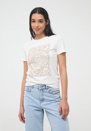 T-shirt bianco in cotone con un dettaglio floreale dorato stampato sul davanti, maniche corte e scollo rotondo. Jeans di denim blu chiaro.