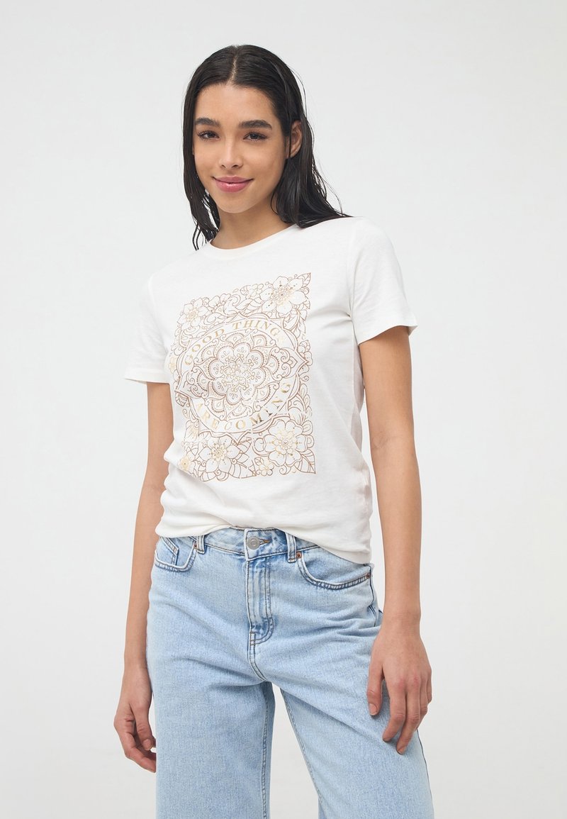T-shirt bianco in cotone con un dettaglio floreale dorato stampato sul davanti, maniche corte e scollo rotondo. Jeans di denim blu chiaro.