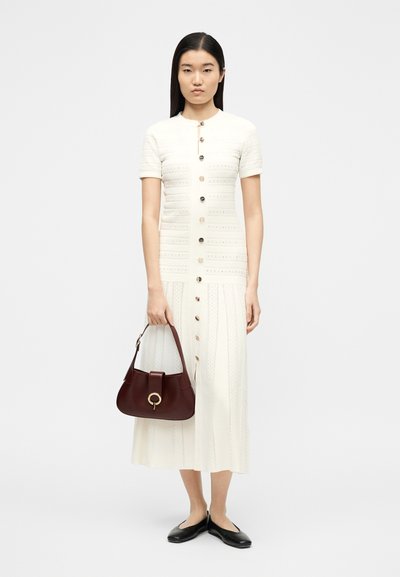 Γυναίκα με λευκό φόρεμα με κοντά μανίκια και κουμπιά, κρατώντας μπορντό τσάντα χειρός, φορώντας μαύρα μπαλαρίνες, στεκόμενη μπροστά σε απλό φόντο.