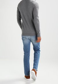 Mann, der einen grauen Pullover, blaue Jeans und braune Sneakers trägt, geht von einem weißen Hintergrund weg.