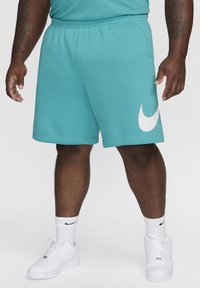 Pantalones cortos Nike en tono verde azulado con un logo blanco, cinturilla elástica y bolsillos laterales. Se llevan con zapatillas blancas y calcetines de caña alta que tienen un logo negro.