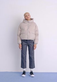 Doudoune beige clair avec capuche, dotée de deux poches avant, associée à un pantalon gris court et des baskets noires. Texture lisse.