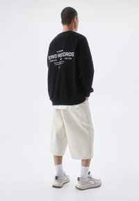 Černá mikina s bílým potiskem na zádech, krémové šortky, bílé ponožky a světle šedé tenisky s akcenty. Jednoduché pozadí.