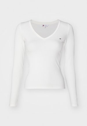 Valkoinen pitkähihainen ribbatun kankaan paita V-pääntiellä, jossa on pieni logo vasemmassa rinnassa, valmistettu kevyestä kankaasta.