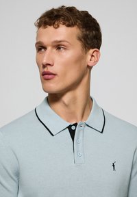 Camisa polo azul claro con un collar y botones negros, que presenta un pequeño logo en el pecho. Hecha de un tejido de punto texturizado.